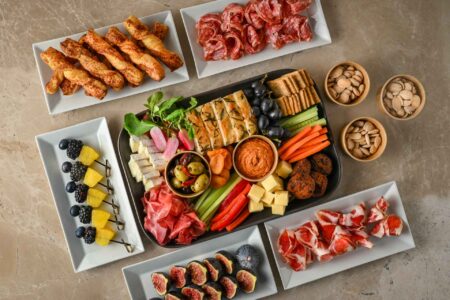 Spread of finger foods with central grazing platter including nuts, figs, fruit skewers, cheese twists, focaccia, vegetable sticks and dips
